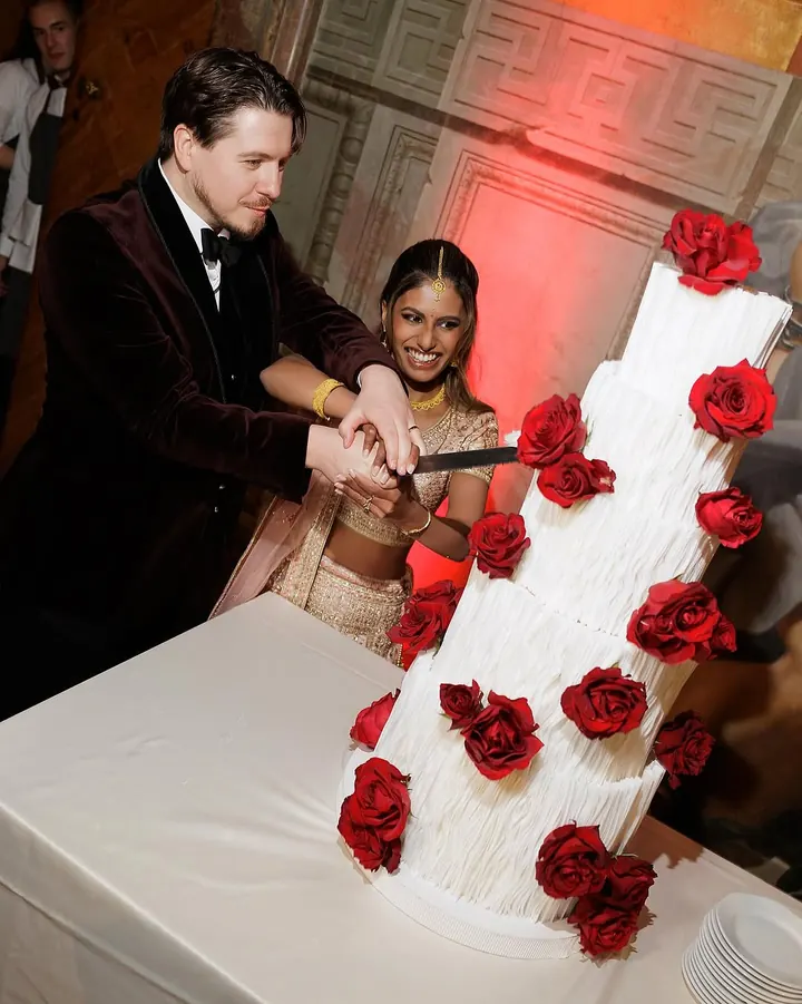 Five-tier white wedding cake covered with fresh red roses, being cut by a couple